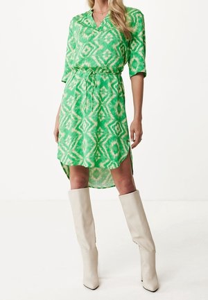 Femme portant une robe verte à motifs longueur genou avec une ceinture nouée à la taille et des bottes blanches hautes jusqu'aux genoux, debout devant un fond uni.