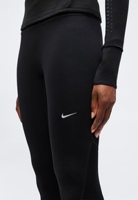 Nærbillede af en person iført sorte Nike leggings og en sort langærmet top med en refleksiv Nike swoosh på låret.