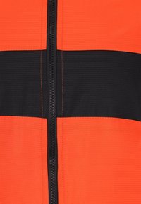 Orange og sort lynlåsjakke med vandrette farveblokke, tekstureret stof og sort syning langs lynlåsen.