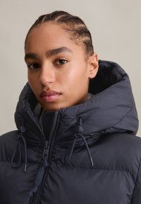 Puffer jacket in dark navy, featuring a high collar, adjustable drawstrings, and double zipper. Smooth fabric with a matte finish.