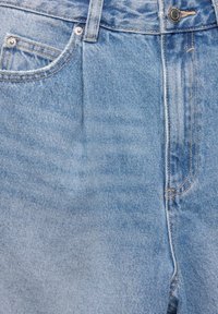 Jean en denim bleu clair en gros plan montrant les poches avant, les passants de ceinture, le bouton et la zone de la fermeture éclair avec des détails de couture visibles.
