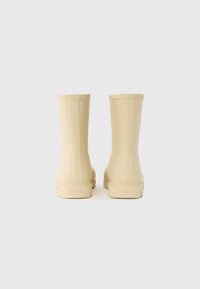 Bottes de pluie en caoutchouc beige avec une texture lisse, une tige haute et une semelle plate. Présente des stries verticales sur les côtés et un logo au niveau du talon.
