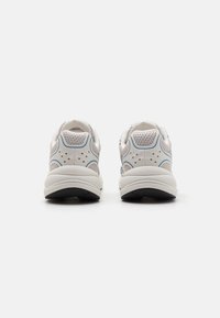 Bílé atletické boty s síťovanými panely a světle modrými akcenty, s zaobleným tvarem a texturovanými gumovými podrážkami. Jsou přítomny perforované detaily.