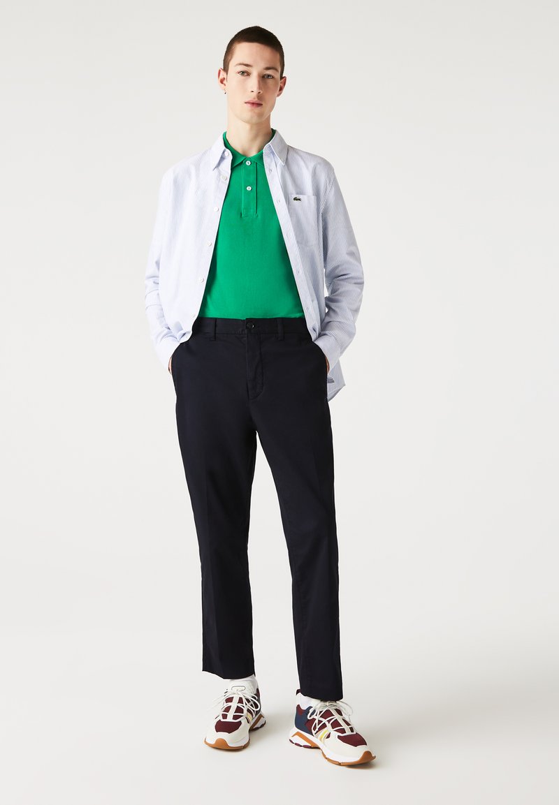 Camiseta polo verde bajo una camisa de botones azul claro a rayas, combinada con pantalones oscuros de color azul marino y zapatillas blancas con acentos burdeos y azul marino.