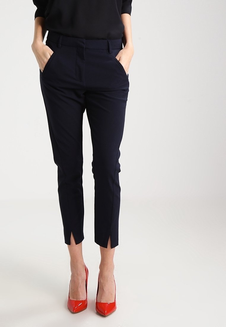 Pantalon bleu marine sur mesure avec poches avant et fentes latérales à l'ourlet, associé à des escarpins rouges brillants. Texture lisse et coupe moderne.