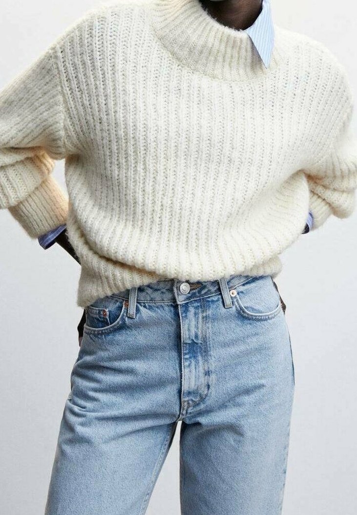 Personne portant un pull en grosse maille blanc par-dessus une chemise bleue à col, rentrée dans un jean taille haute bleu clair, les mains sur les hanches.