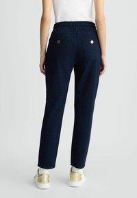 Pantalons bleu marine avec une coupe décontractée, une taille élastique, deux poches arrière avec des accessoires en or, et un matériau texturé, légèrement scintillant.