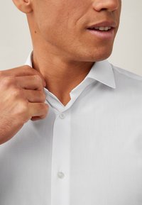 Moški prilagaja ovratnik bele srajce z gumbi, izrezano na bradi in roki, nevtralno ozadje.