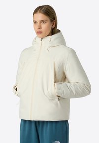 Kvinna i en krämfärgad huvtröja med dragkedja och händerna i fickorna, iförd blåa The North Face-byxor, som tittar åt sidan.
