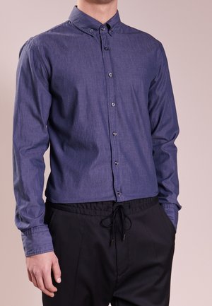 Mężczyzna w granatowej koszuli z długim rękawem na guziki i czarnych spodniach z sznurkiem, stojący z jedną ręką w kieszeni na jednolitym tle.