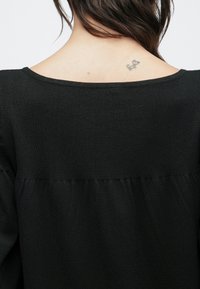 Μαύρο πλεκτό τοπ με φαρδιά λαιμόκοψη και υφασμάτινη επιφάνεια στο σώμα, με οριζόντια ραφή κάτω από τους ώμους. Είναι ορατό ένα μ�ικρό τατουάζ.