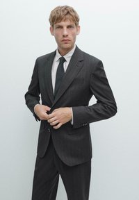 Dunkelgrauer Nadelstreifenanzug mit einem einreihigen Blazer und passender Hose. Enthält ein weißes Hemd und eine schwarze Krawatte, glatte Stofftextur.
