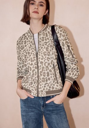 Jonge vrouw met een beige leopard print jas met rits, blauwe spijkerbroek, draagt een zwarte schoudertas, staand met handen in haar zakken.