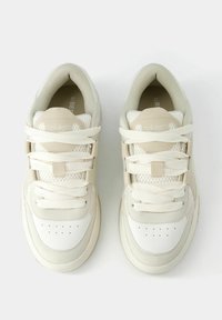 Ett par beigefärgade och vita sneakers med meshtyg och tjocka vita snören, vy från ovan på en vit bakgrund.