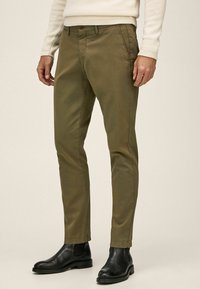 Khakifarbene Chinos mit geradem Schnitt aus Baumwolle, ausgestattet mit schlanken Taschen, glatter Stoffstruktur und kombiniert mit schwarzen Schnürstiefeln.