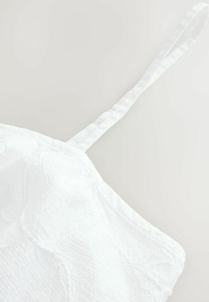 Balta auduma blūze ar rakstainu faktūru, ar plānu regulējamu siksniņu un maigu, nedaudz krunkotu izskatu. Redzama nav nekāda aparatūra.