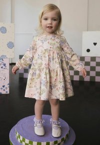 Lyserød kjole med blomstermønster, lange ærmer og lagdelt design. Kombineret med lyse lilla sko og flæsekantede ankelstrømper på en rund piedestal.