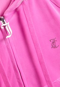 Prim-plan al unei jachete din fleece roz aprins cu fermoar, un pandantiv argintiu pentru fermoar în formă de litera „J” și un mic logo „JC” cu ținte.