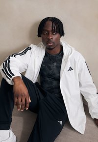 Casaco de vento branco com riscas pretas, camisa preta texturada e calças pretas. Logótipo da Adidas em destaque e fundo neutro.