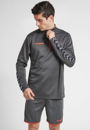 Mężczyzna w szarej odzieży sportowej z czarnymi paskami w kształcie chevron na rękawach zapina górę, nosi dopasowane spodenki z pomarańczowym wykończeniem i logo.