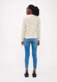 Šviesiai rudos megztinis su deimantiniu raštu, ilgomis rankovėmis ir rausvais kraštais, derinamas su šviesiai mėlynomis džinsomis ir juodais platforminiais batais.