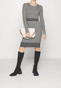 Grau gestreiftes Strickkleid mit langen Ärmeln, schwarzer gestreifter Taillengürtel, kombiniert mit einer weißen gesteppten Tasche und schwarzen kniehohen Stiefeln mit Absatz.