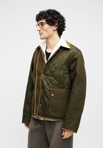 Νέος άνδρας με σγουρά μαλλιά και γυαλιά που φοράει μια χακί μπουφάν με μεγάλο καπιτονέ και μεγάλες τσέπες, ένα λευκό πουκάμισο με γιακά και γκρι παντελόνι.