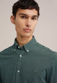 Camisa de botones verde oscuro con una textura en la tela, cuello puntiagudo y botones blancos. Presenta un pequeño emblema bordado en el pecho.