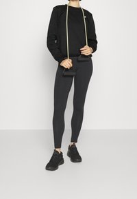 Felpa nera con collo rotondo, leggings neri e scarpe da ginnastica nere. Tiene in mano una banda di resistenza verde con manici.
