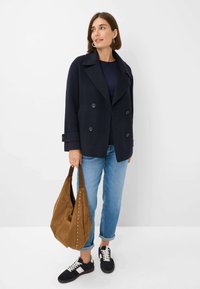 Manteau officier bleu marine à double boutonnage, porté sur un haut sombre, associé à un jean bleu clair, un sac à bandoulière marron avec des clous, et des baskets noires.