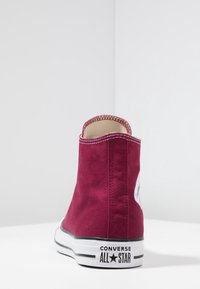 Bordowe trampki Converse wysokie, wykonane z materiału płóciennego, z białą gumową podeszwą, kontrastującym czarnym paskiem i okrągłym logo po boku.