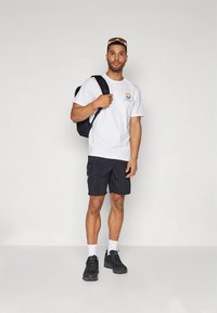 Hvit t-skjorte med en fargerik bølgedesign, svarte cargoshorts, hvite ankelsokker og svarte joggesko. Modellen bærer en svart ryggsekk.