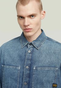 Ung man med kort blont hår som bär en blå denimskjorta med knappar, framfickor och en G-Star Originals-etikett.