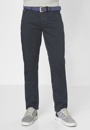 Dark gray slim-fit pants with visible stitching, paired with a patterned blue belt. Feet in gray sneakers with textured mesh accents.