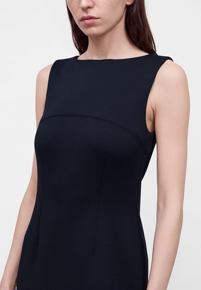 Femme portant une robe noire sans manches avec un col droit et haut sur un fond clair uni.