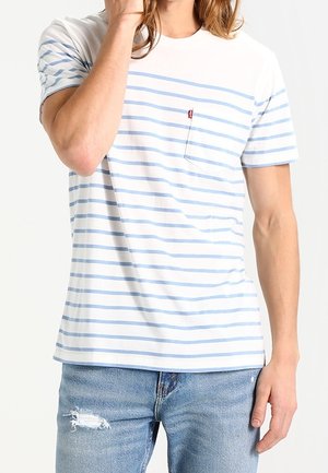 Jeune personne portant un t-shirt à manches courtes rayé blanc et bleu clair et un jean bleu usé, avec une main proche du visage.