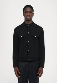 Černá button-up bunda s dvěma náprsními kapsami, stříbrným kováním a strukturovaným střihem. Vyrobeno z hladké, texturované tkaniny.