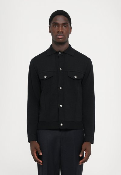 Ung mand iført en sort knapjakke med brystlommer og mørke bukser, stående mod en ensfarvet hvid baggrund.