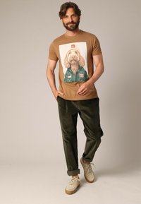 Bruine graphic t-shirt met een hondenprint, gecombineerd met groene katoenbroeken en beige sneakers met bruine zolen. Casual outfitontwerp.