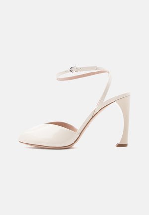 Λευκό παπούτσι με ψηλό τακούνι από λουστρινι, μυτερή μύτη και λεπτό λουράκι στον αστράγαλο με ασημένια αγκράφα.