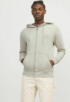 Lys grå zip-up hoodie med snor, kengurulommer og kontrasterende syninger. Modellen bærer beige bukser med gyldne accenter.