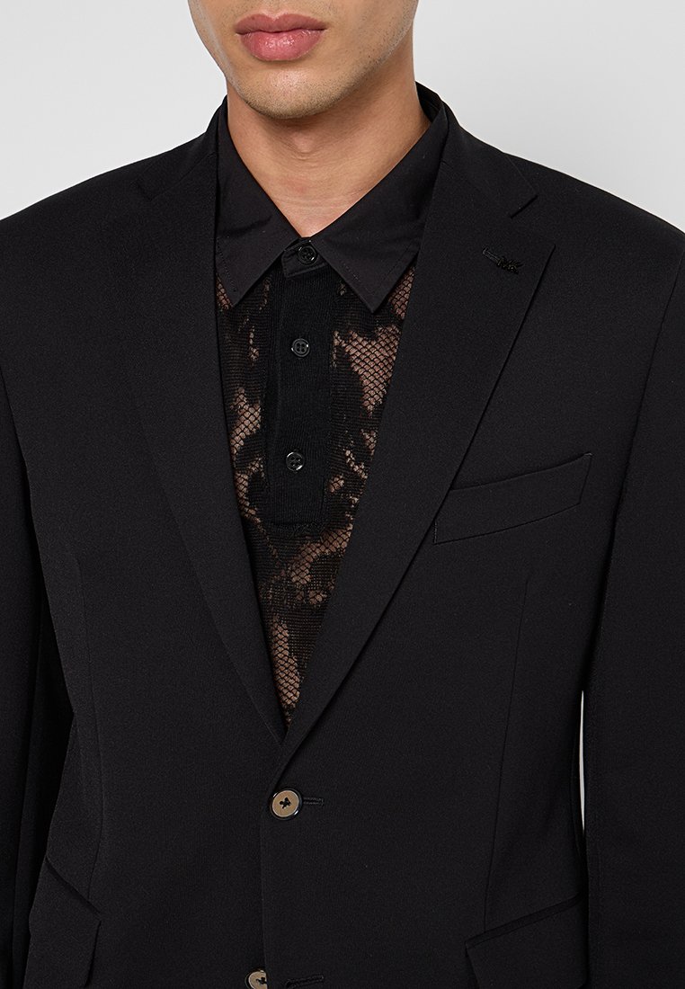 Un giovane indossa un blazer nero sopra una camicia a rete con stampa, mostrando una scelta di moda moderna e audace. Lo sfondo è neutro.