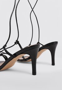 Sandalias de tacón alto negras con tiras, acabados texturizados, cordones delgados y plantilla beige. Presentan un diseño elegante de punta afilada.