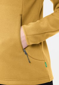 Geel fleecejack met een rits zijzak en verstelbare trekkoorden. Gladde textuur en aangesloten mouwen. Groene merketiket aan de onderzijde.