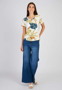 T-shirt à manches courtes, couleur crème, avec un imprimé tropical bleu et orange ; associé à un jean en denim bleu à jambes larges et des sandales à talons blocs blanches.