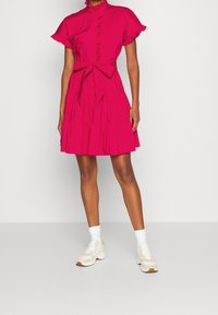 Kvinna i en ljus rosa kortärmad klänning med volanger och ett knytbälte i midjan, tillsammans med vita strumpor och beige sneakers.