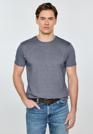 Jauns vīrietis, tērpies pelēkā īsroku t-kreklā un zilās džinsās ar brūnu jostu, stāvot ar vienu roku kabatā uz vienmērīga fona.