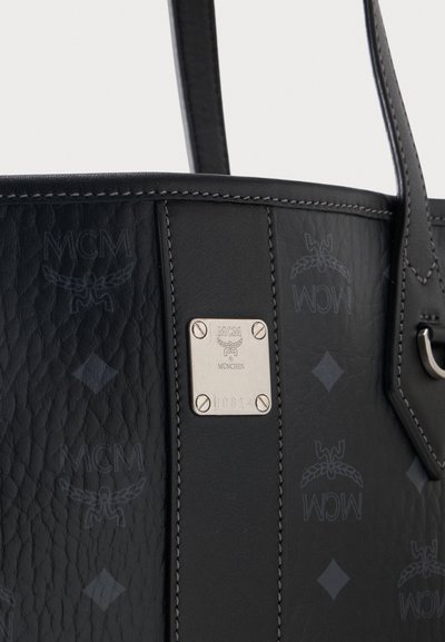 Μαύρη δερμάτινη τσάντα MCM με ανάγλυφο μοτίβο λογότυπου, κάθετη κεντρική ρίγα και ασημένια μεταλλική πινακίδα λογότυπου με βίδες και σειριακό αριθμό.