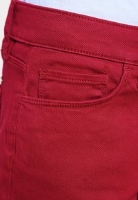 Jeans de mezclilla rojos que presentan un bolsillo trasero estándar, detalles de costura visibles y una textura suave. La cintura se muestra en la parte superior.