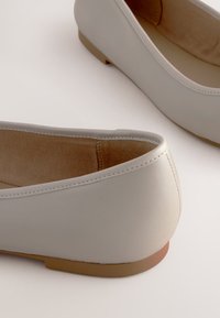 Gros plan de deux ballerines en cuir blanc cassé avec une doublure intérieure couleur cuir clair et des semelles marron clair sur une surface blanche.